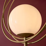 Close-up of the Endon Brackshaw floor lamp showing its opal glass orb cradled within three interlinking brushed gold rings against a deep burgundy wall.