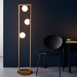 Cranford 3 Light Floor Lamp in brushed gold, tall rectangular frame with three glowing opal glass spheres, shown beside a black chair and desk.