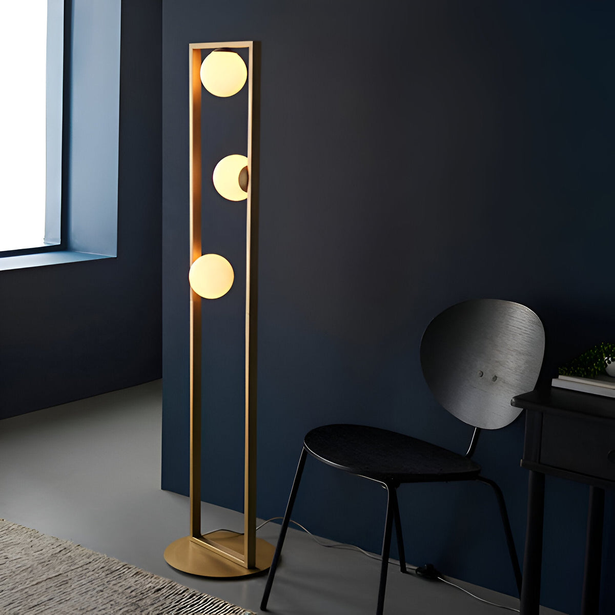 Cranford 3 Light Floor Lamp in brushed gold with three opal glass globes on a tall angular frame, shown beside a black chair in a dark-walled room.