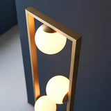 Cranford 3 Light Floor Lamp in brushed gold, angular metal frame holding three glowing opal glass spheres against a dark wall.