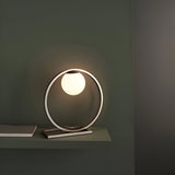 Brackstead Table Lamp in brushed silver, a circular metal frame on a rectangular base holding a glowing opal glass sphere, shown lit on a shelf.