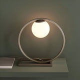 Brackstead Table Lamp in brushed silver, a circular metal ring on a rectangular base holding a glowing opal glass sphere, shown on a shelf.