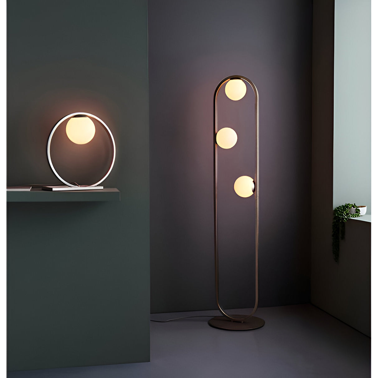 Brackstead Table Lamp in brushed silver, a ring-shaped frame holding a glowing opal glass sphere, shown lit on a shelf in a modern living room.