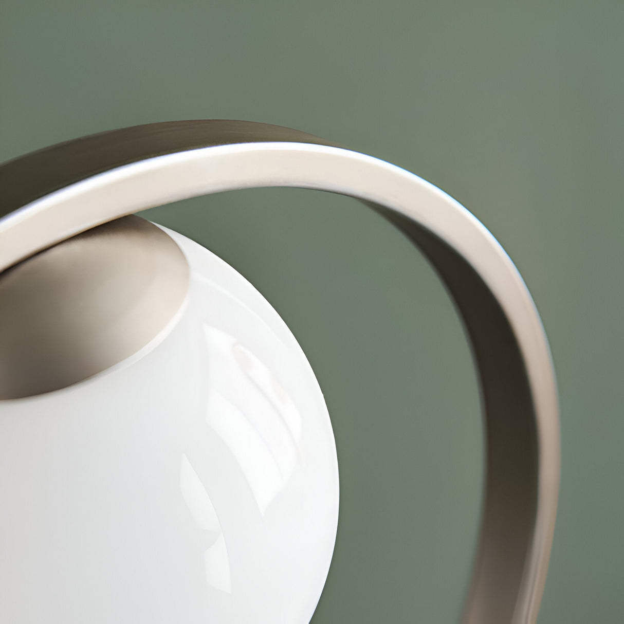Close-up of the Brackstead Table Lamp's brushed silver circular frame curving beside its glossy opal glass sphere, set against a sage green background.