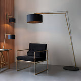 Endon Carlfield cantilevered floor lamp in matt brass with a black cotton drum shade, beside a black and brass armchair in a modern interior.