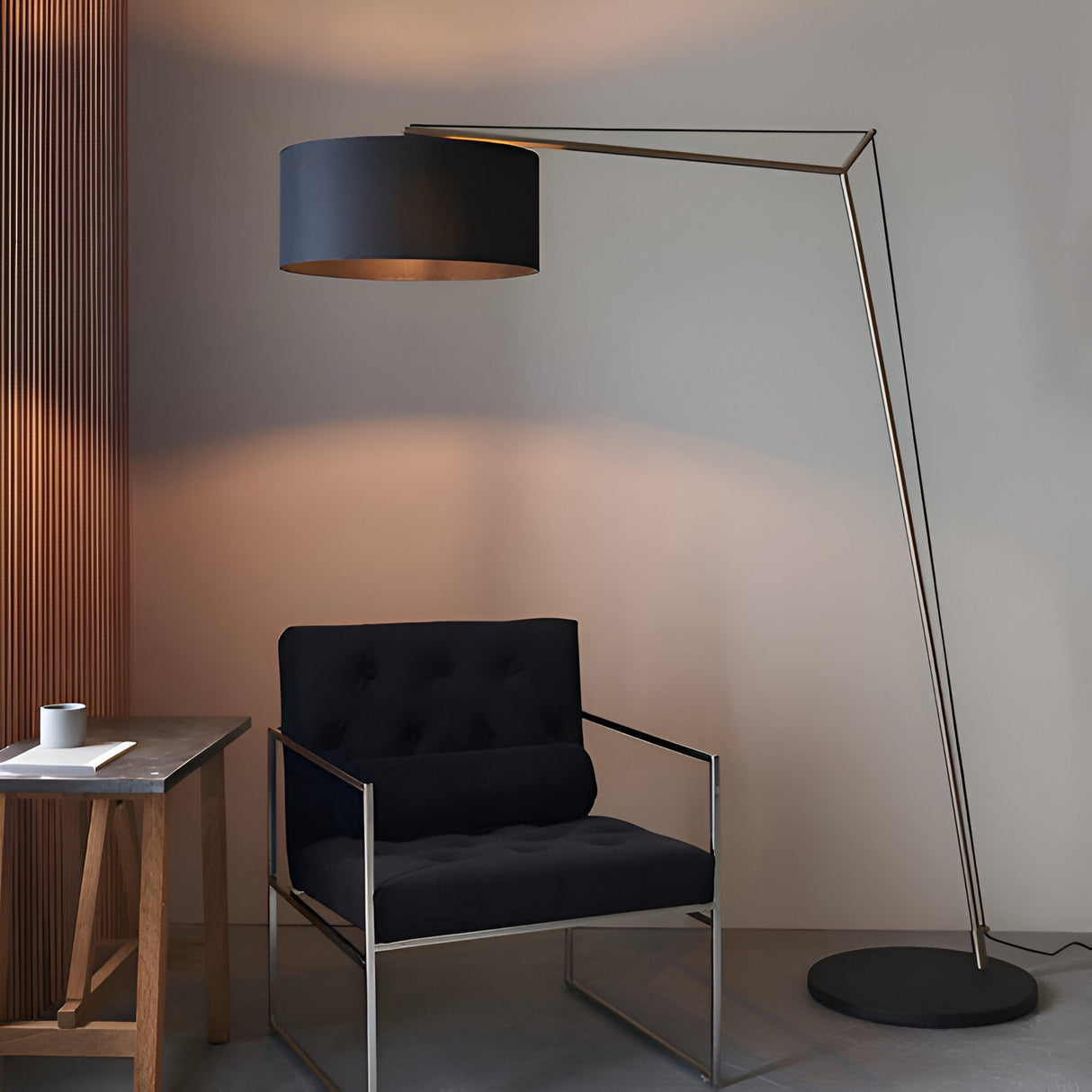 Carlfield Floor Lamp in matt nickel with a cantilevered arm and black cotton drum shade, standing beside a black armchair on a matt black round base.