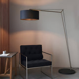 Carlfield Floor Lamp in matt nickel with a cantilevered arm and black cotton drum shade, standing beside a black armchair on a matt black round base.