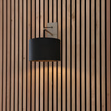 Carlfield Wall Light in nickel with a black rectangular base, cantilevered stem, and black fabric drum shade, mounted on a wood slat wall.