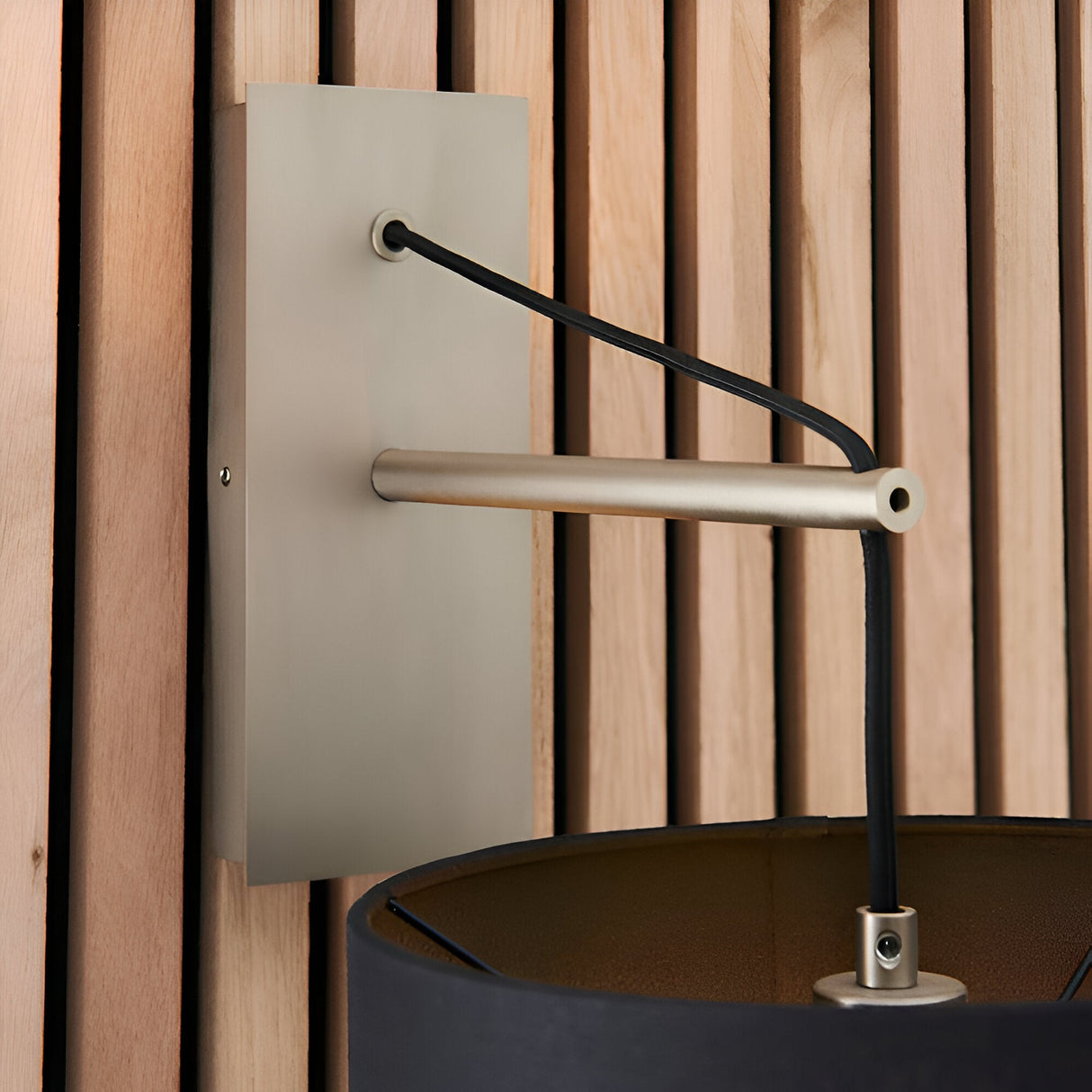 Carlfield Wall Light in Nickel, showing the matt nickel rectangular backplate, cantilevered stem, black fabric cord and shade, against a wooden slatted wall.
