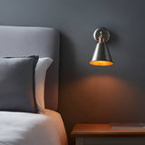 Endon Brackley wall light in aged copper and pewter with adjustable conical shade, illuminated beside a grey upholstered bed and wooden nightstand.