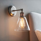 Brackley glass wall light in pewter and copper finish with cone-shaped clear glass shade and exposed bulb, mounted above a bed headboard.