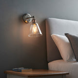 Brackley Glass Wall Light in pewter and copper, adjustable cone shade with visible bulb, mounted above a bed with grey headboard.