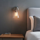 Brackley Glass Wall Light in pewter and copper, cone-shaped clear glass shade with visible bulb, mounted above a bedside table beside a grey headboard.