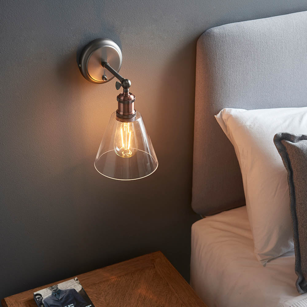 Brackley Glass Wall Light in pewter and copper with adjustable head and clear conical shade, shown lit above a bed headboard.