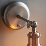 Close-up of the Brackley Glass Wall Light in pewter and copper, showing the round wall plate, adjustable ball-joint arm, and knurled copper bulb holder.
