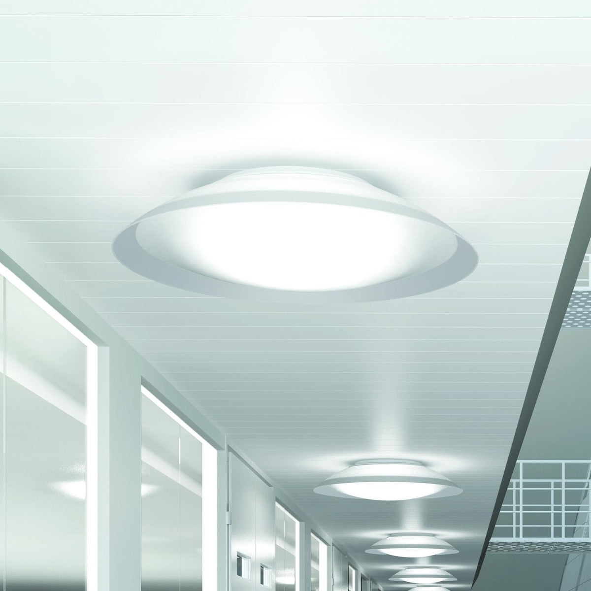 Lens LED Flush Ceiling Light in white, round domed diffuser with flared rim, shown glowing on a hallway ceiling alongside matching fixtures receding down the corridor.