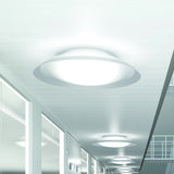 Lens LED Flush Ceiling Light in white, round domed diffuser with flared rim, shown glowing on a hallway ceiling alongside matching fixtures receding down the corridor.