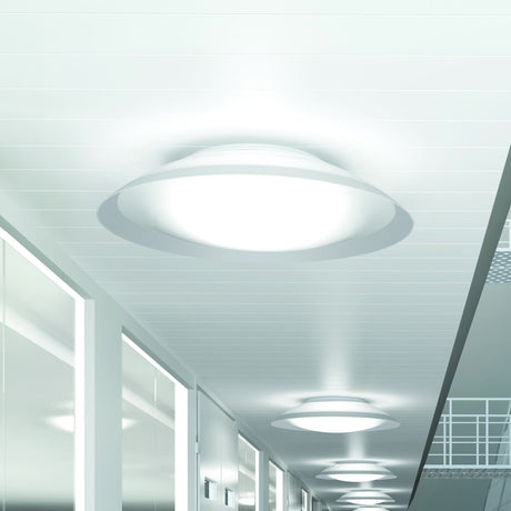 Lens LED Flush Ceiling Light in white, round domed diffuser with flared rim, shown glowing on a hallway ceiling alongside matching fixtures receding down the corridor.