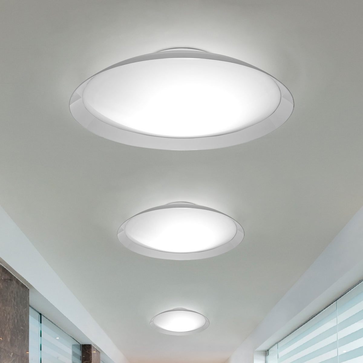 Lens LED Flush Ceiling Light in white shown in three sizes along a hallway ceiling, each with a round acrylic diffuser in a flared iron rim casting a soft glow.