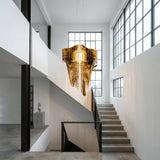 A modern, minimalist interior featuring concrete stairs, large industrial windows, white walls, and the Aeris 5 Light Large Chandelier in gold casting ambient light over the stairwell. Natural light flows through the space.