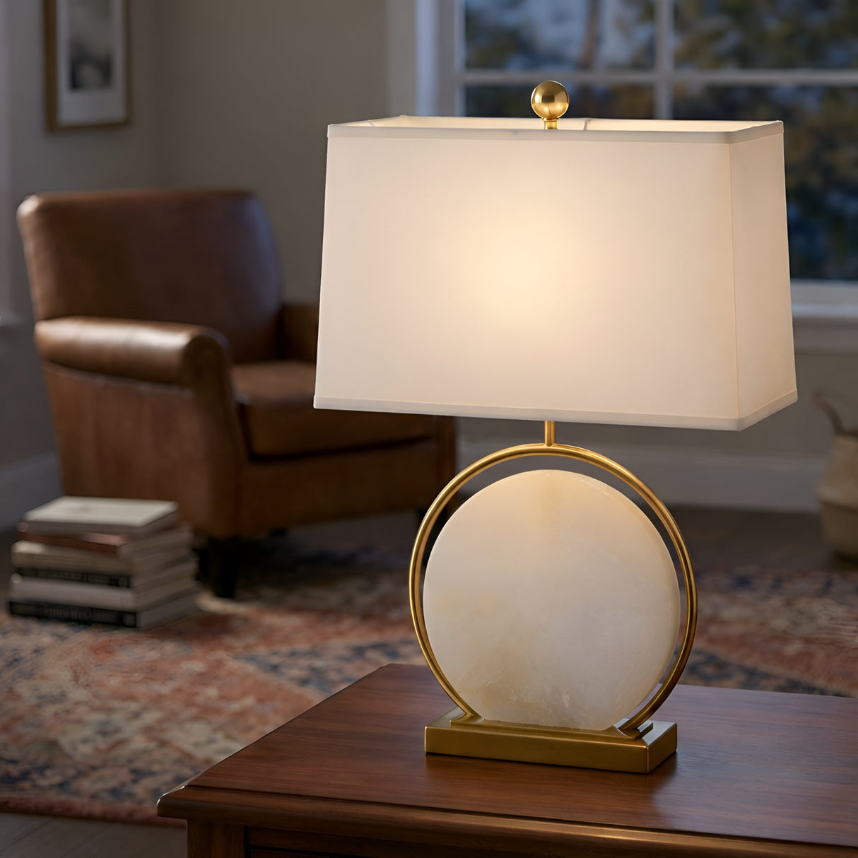 The Orivane 1 Light Circular Alabaster Table Lamp in brass and soft white rests on a wooden table, with a brown leather armchair, books, and a patterned rug visible near the window in the background.