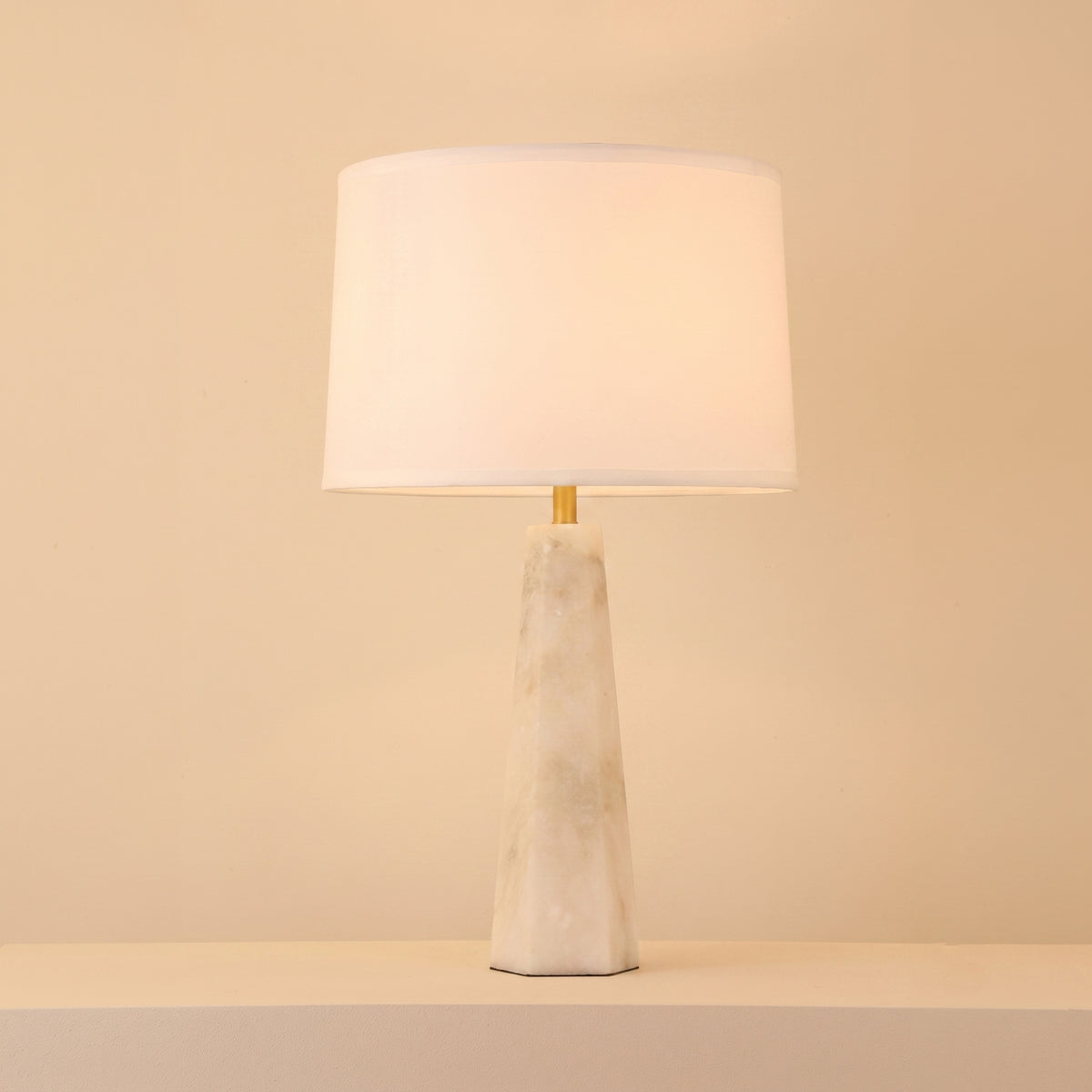 Orix Alabaster Pillar Table Lamp with tapered faceted white alabaster column, brushed brass collar, and soft white drum shade on a plain light background.