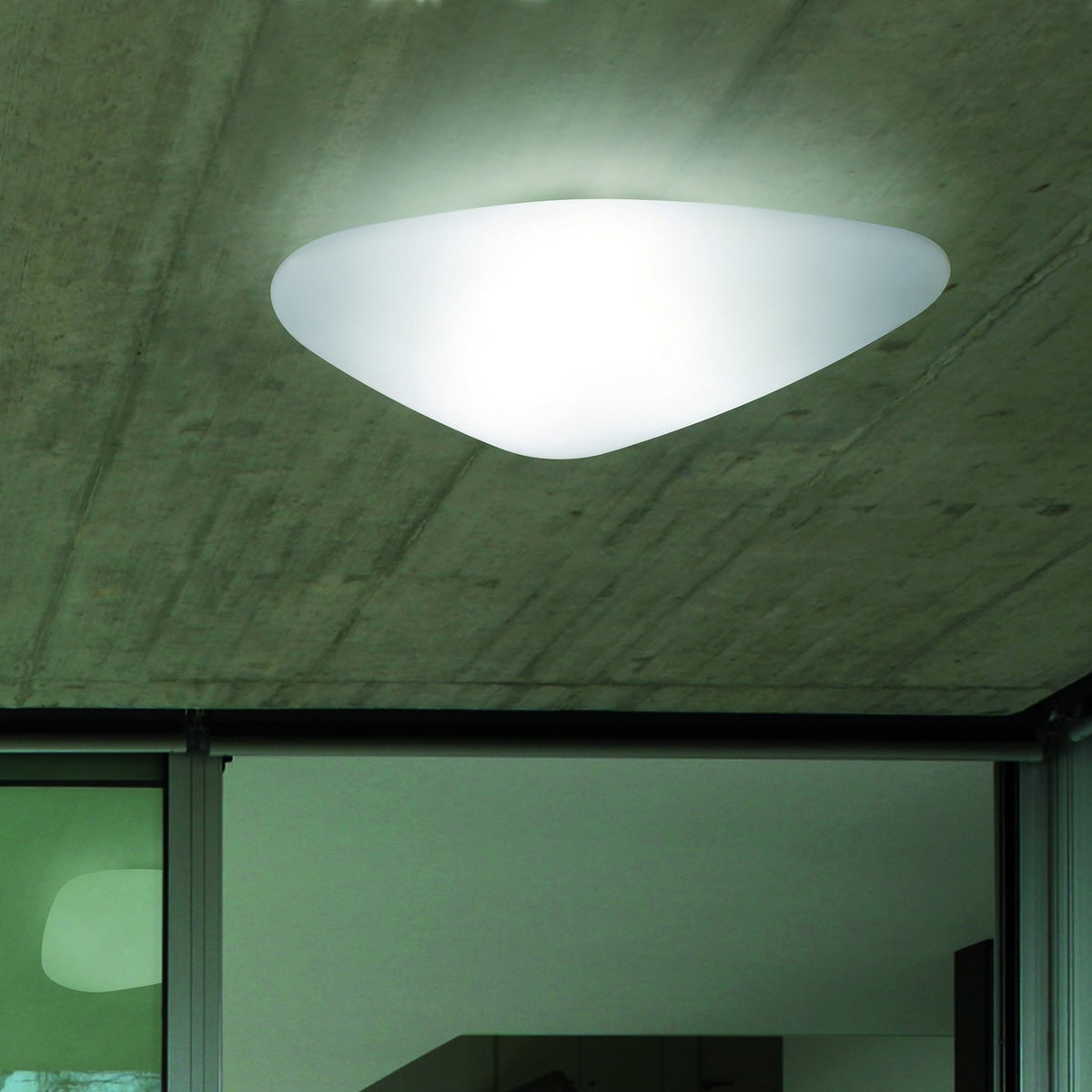 Stone LED Flush Ceiling Light in white, an asymmetric pebble-shaped diffuser glowing on an exposed concrete ceiling above a glazed interior doorway.