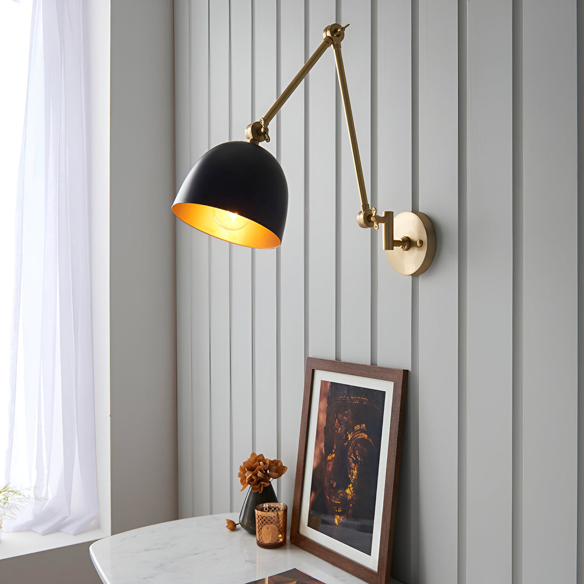 Caldhurst Wall Light in brass and black with an adjustable jointed arm and dome shade, mounted above a marble console table with framed art.