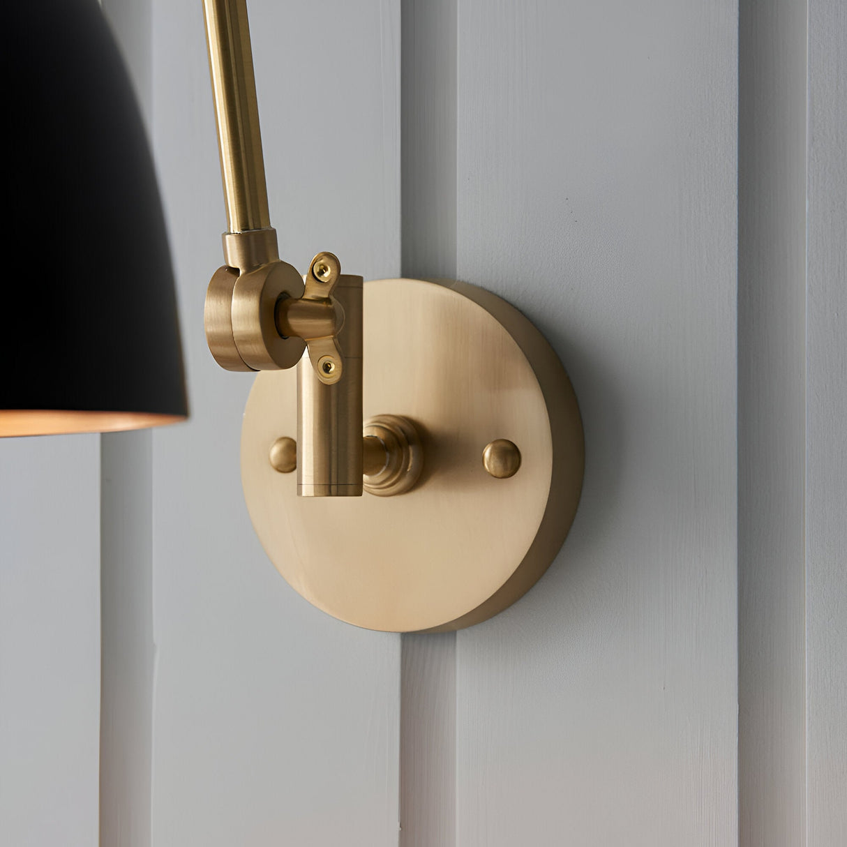 Caldhurst Wall Light in brass with adjustable arm, round wall plate and knurled joint detail, paired with a matt black shade, mounted on a grey panelled wall.