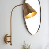 Chartmere Wall Light in antique brass with curved arm and angled cone shade, mounted beside an oval mirror on a light grey wall.