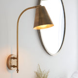 Chartmere Wall Light in antique brass with tall curved arm and angled cone shade, mounted beside an oval mirror on a white wall.