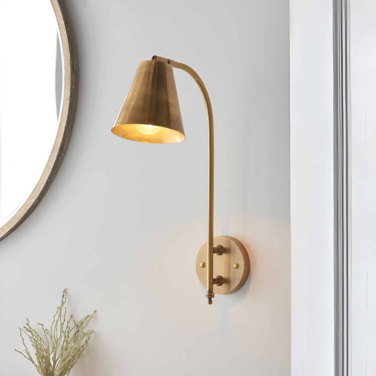 Chartmere Wall Light in antique brass with a curved arm and cone-shaped directional shade, mounted on a light grey wall beside a round mirror.