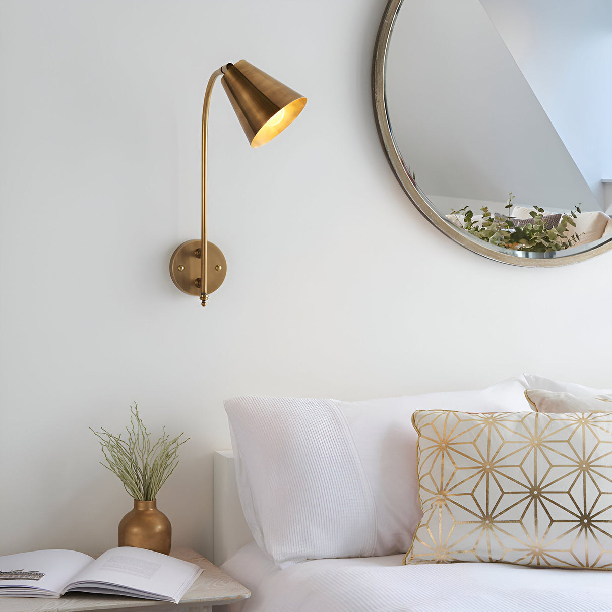 Chartmere Wall Light in antique brass mounted above a bedside table, showing its adjustable cone shade and long arm in a bedroom setting near a round mirror.