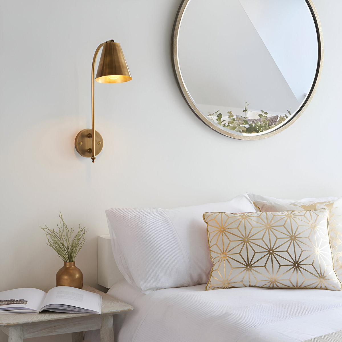 Chartmere Wall Light in antique brass with an adjustable, angled cone shade, mounted above a bedside table beside a round mirror in a bedroom setting.