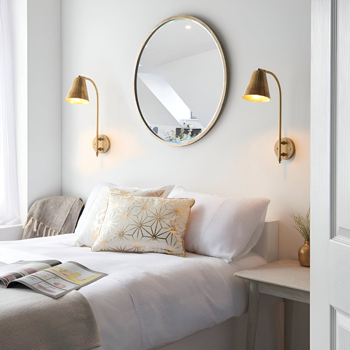 Pair of Chartmere antique brass wall lights with angled cone shades flanking a round mirror above a bedroom headboard.