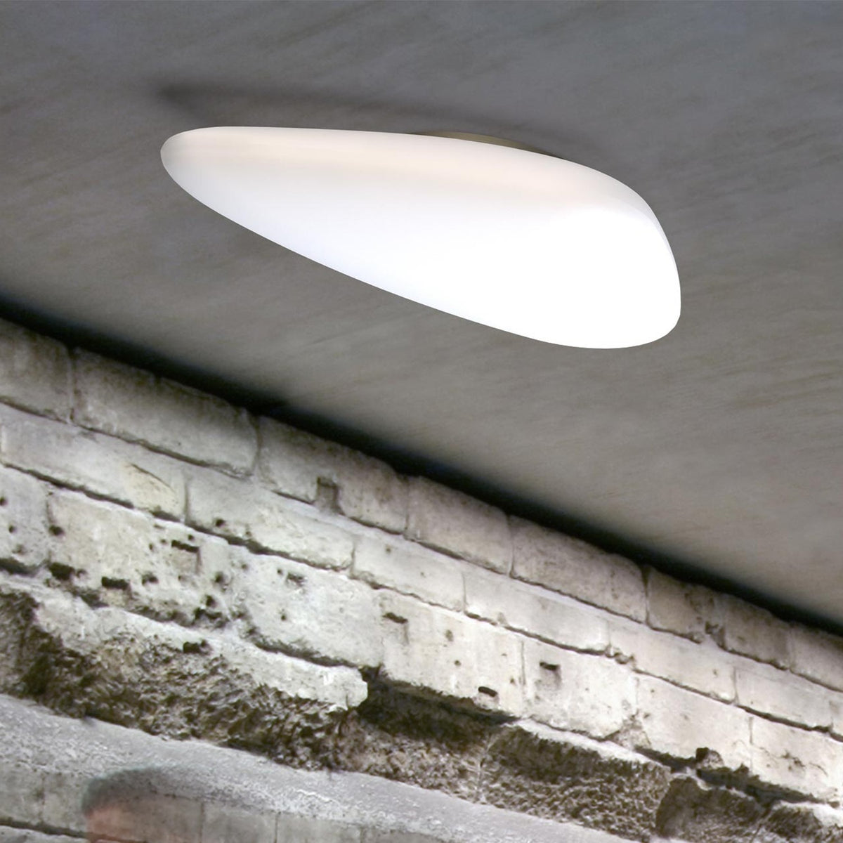 Stone LED Outdoor Flush Ceiling Light in white, pebble-shaped diffuser mounted flush against a grey ceiling above an exposed brick wall, glowing evenly with no visible frame.