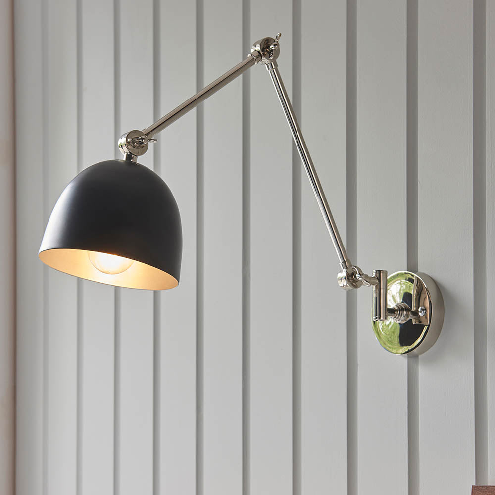 Endon Caldhurst adjustable wall light with polished nickel articulated arm, matt black dome shade and lit E27 bulb, mounted on grey panelled wall.
