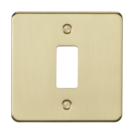 The 1 Gang Grid Faceplate - Brushed Brass boasts a premium grade steel flat plate with a brushed brass finish and a rectangular opening for the switch toggle. Visible screws at the top and bottom provide secure mounting.