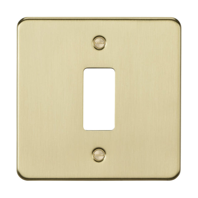 The 1 Gang Grid Faceplate - Brushed Brass boasts a premium grade steel flat plate with a brushed brass finish and a rectangular opening for the switch toggle. Visible screws at the top and bottom provide secure mounting.