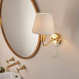 Endon Bevton bathroom wall light in satin brass with a curved arm, ivory fabric shade and pull cord, mounted beside an oval brass mirror above a basin.