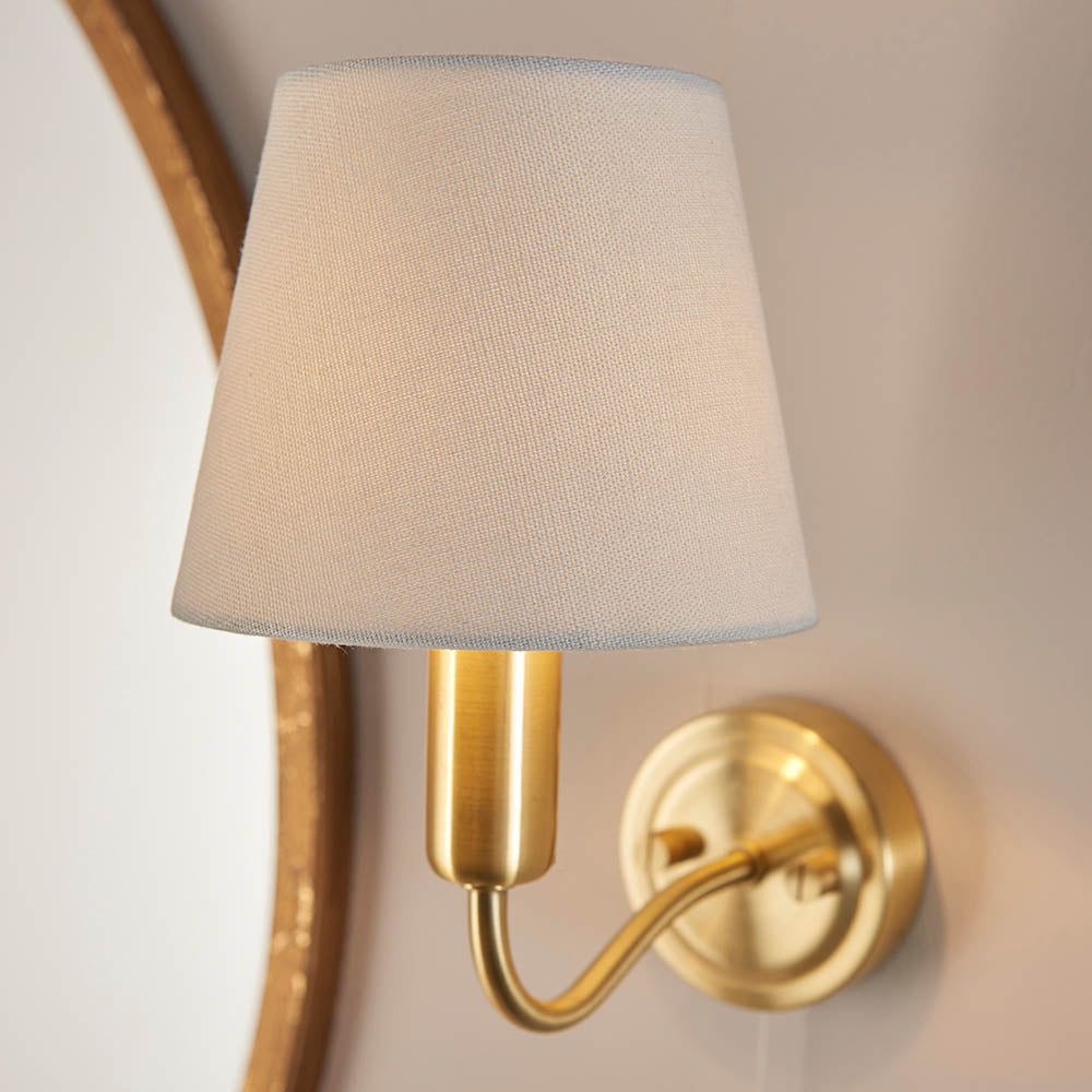 Endon Bevton bathroom wall light with a curved satin brass arm and tapered ivory fabric shade, mounted beside a round wooden-framed mirror.