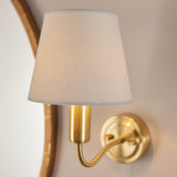 Endon Bevton bathroom wall light with a curved satin brass arm and tapered ivory fabric shade, mounted beside a round wooden-framed mirror.