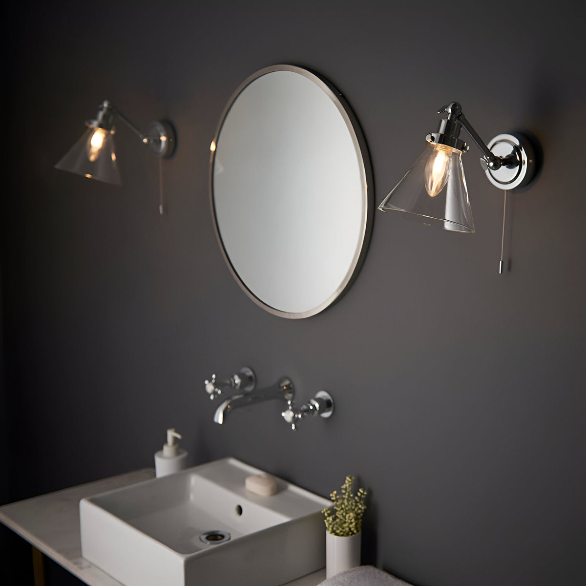 Endon Brackwood chrome bathroom wall lights with clear glass cone shades and pull cords, flanking a round mirror above a white basin on a dark grey wall.