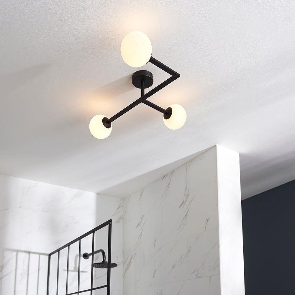 Endon Kelham 3 Light semi-flush bathroom ceiling light in black with three opal glass globes on angular arms, mounted to a white ceiling above marble tiling.