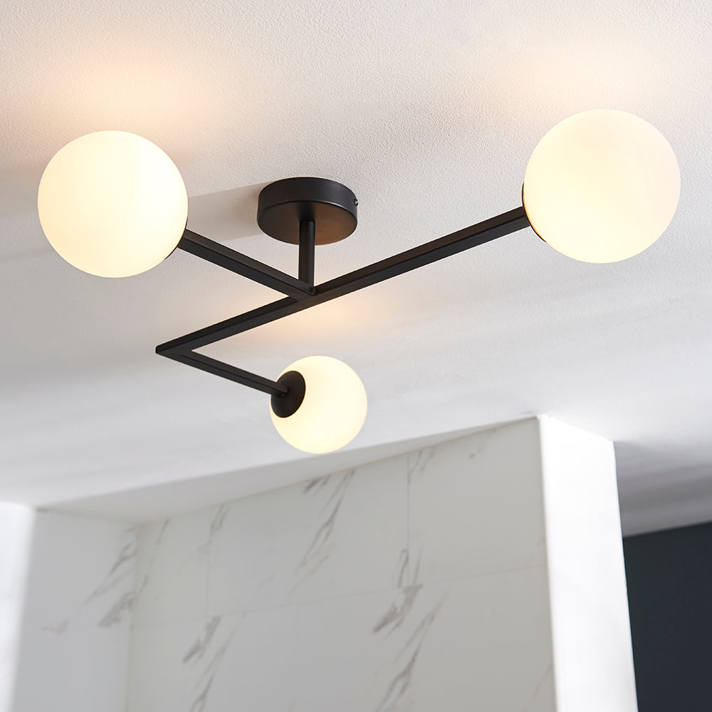 Endon Kelham 3 light semi-flush bathroom ceiling light in black, with three white glass globe shades on angular arms fixed to a white ceiling.