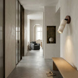 Minimalist hallway with light gray walls and floors, a wooden bench, slippers, modern lighting featuring the Korvane 1 Light Travertine Spotlight in Walnut Brown & Light Beige, a ceramic vase in a niche, and a view into a bright living area with an armchair.