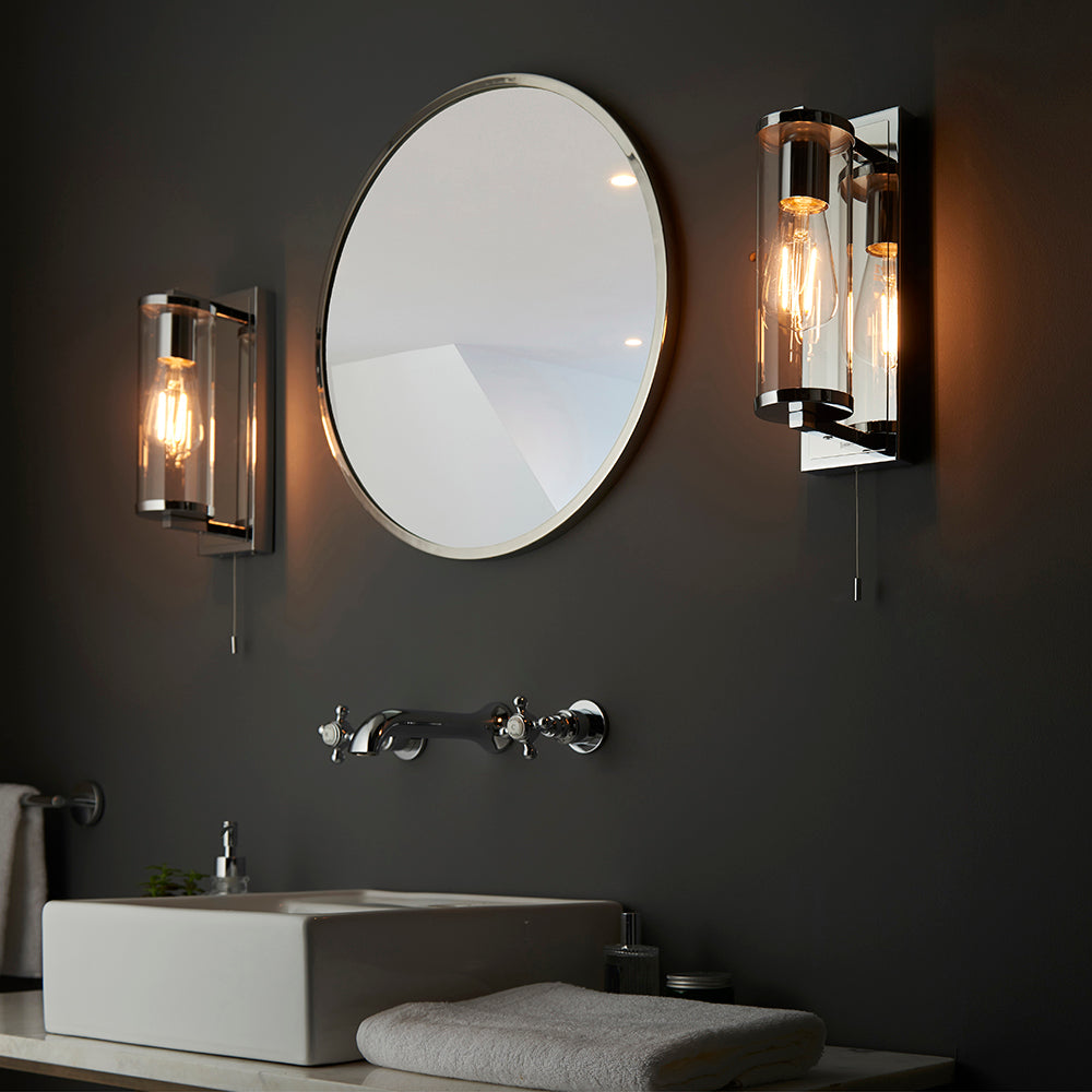 Endon Cranfield bathroom wall lights in chrome with clear glass tube shades and pull cords, flanking a round mirror above a basin on a dark grey wall.