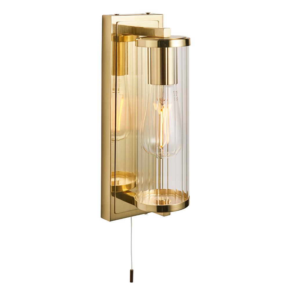 Endon Cranfield IP44 bathroom wall light in brass with ribbed clear glass tube shade, E27 filament bulb and pull cord switch on a plain white background.