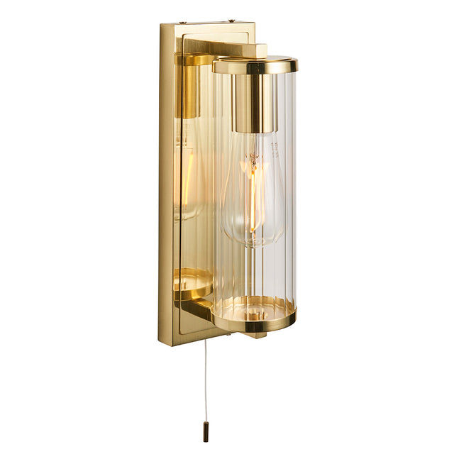 Endon Cranfield IP44 bathroom wall light in brass with ribbed clear glass tube shade, E27 filament bulb and pull cord switch on a plain white background.