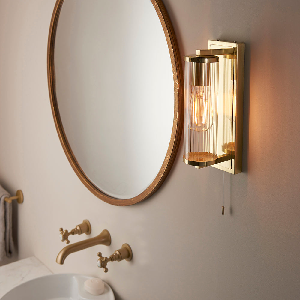 Endon Cranfield brass bathroom wall light with ribbed clear glass tube shade and pull cord, mounted beside a round wood-framed mirror above a brass tap.